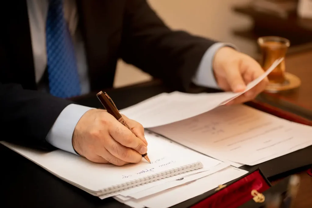 A professional lawyer writing on a notepad with a pen, reviewing documents on a desk cluttered with papers, alongside a cup of tea in the background, lemonlawassist