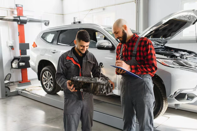 Two mechanics in a car repair workshop, one holding a car part and the other reviewing notes on a clipboard, standing near an SUV with its hood open, lemonlawassist