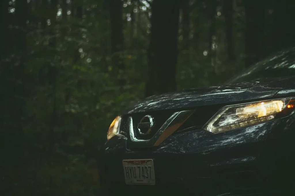 Close-up of a Nissan car with its headlights on, parked in a dimly lit forest.