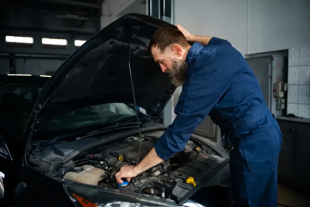 Mechanic in blue overalls checking under the hood of a car in an auto repair shop, lemonlawassist