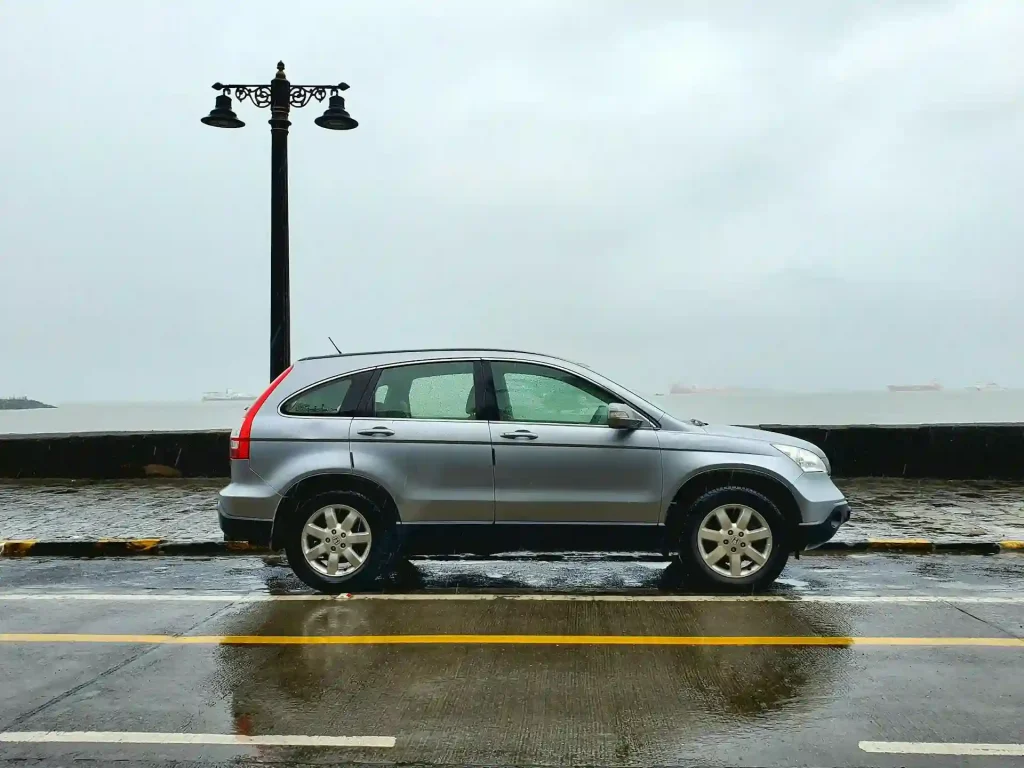 Side view of a silver SUV parked on a wet road near the coast, with a lamp post in the background on a rainy day, lemonlawassist