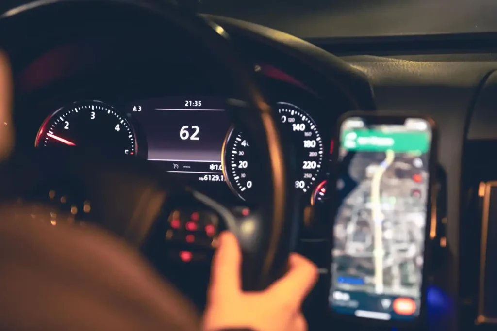 Close-up of a car dashboard at night, showing a speedometer at 62 mph and a smartphone mounted, lemonlawassist
