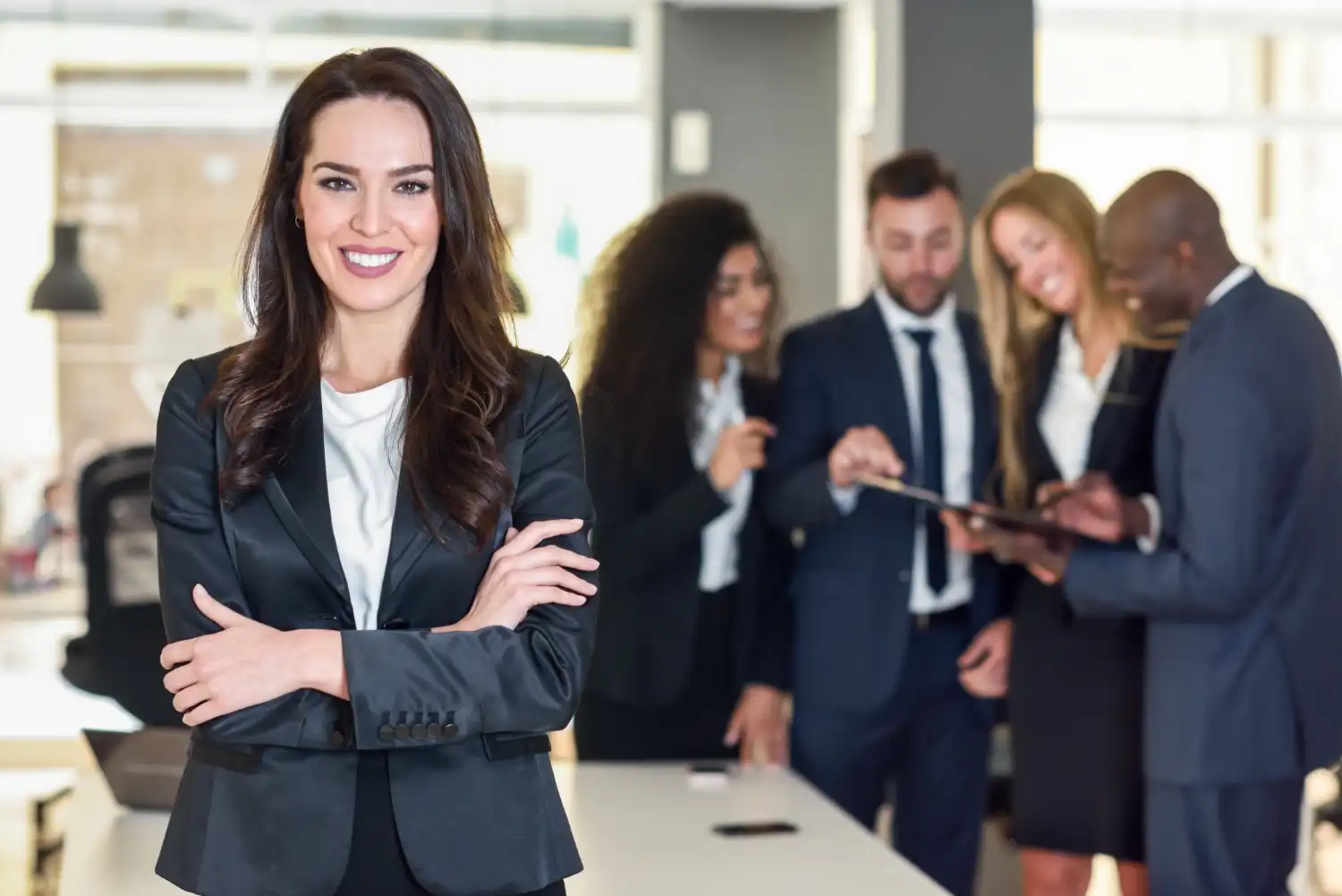 Confident attorney in a formal suit standing with arms crossed in the foreground, while her diverse team collaborates in the background, lemonlawassist