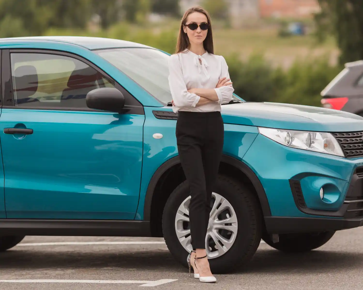 Confident woman in sunglasses standing beside a blue SUV in a parking lot, arms crossed, wearing a white blouse and black pants, lemonlawassist
