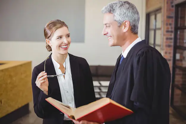 Image of two legal professionals in conversation, with a male lawyer wearing a black robe holding an open book, and a smiling female colleague holding glasses, lemonlawassist