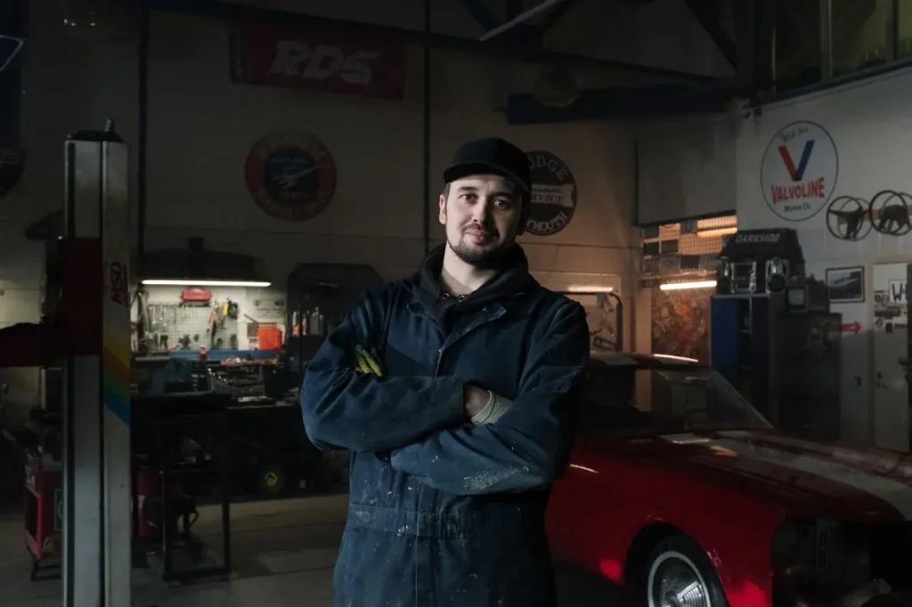 Man in blue zip up jacket standing near car repair shop, lemonlawassist