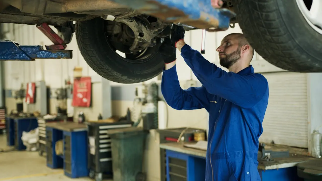 Mechanic in a blue coverall inspecting and repairing a vehicle’s undercarriage in a professional auto repair shop, lemonlawassist