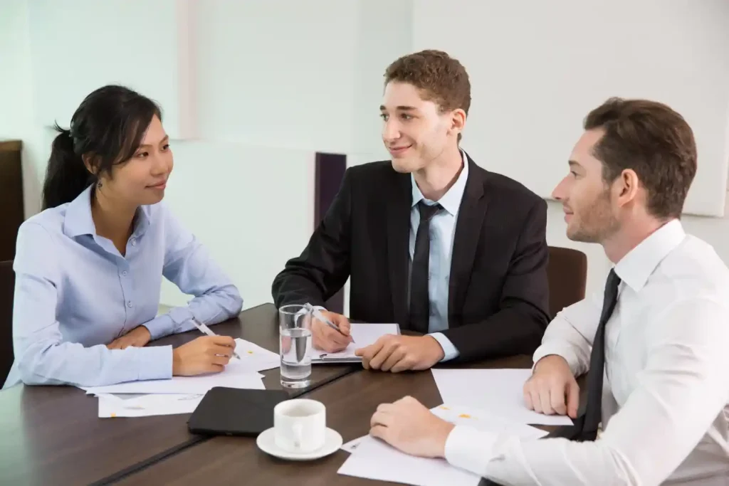 Three business professionals in a meeting, discussing documents around a conference table in an office setting, lemonlawassist