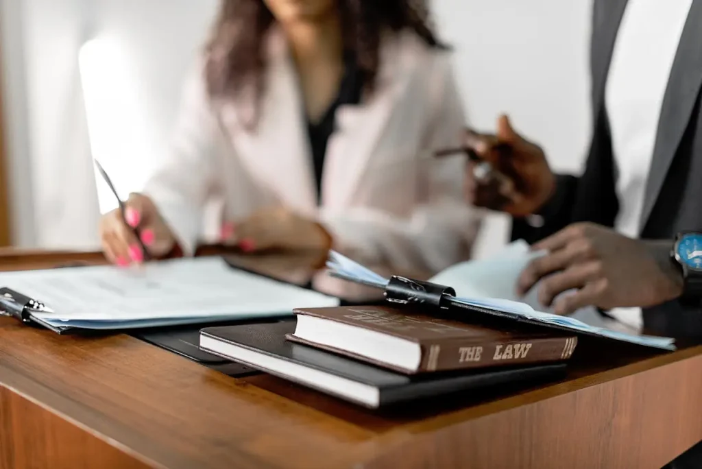 Two individuals discussing legal documents at a desk, with a law book and paperwork prominently placed in the foreground, lemonlawassist