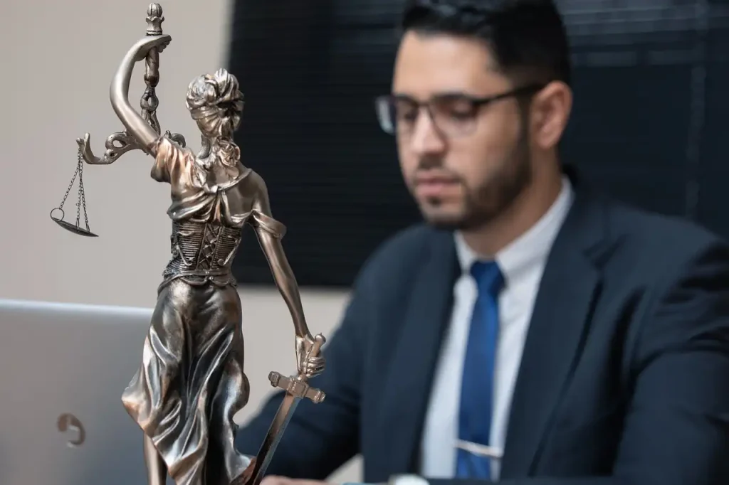 Bronze statue of Lady Justice holding scales and a sword with a male lawyer in a suit and glasses working in the background, lemonlawassist