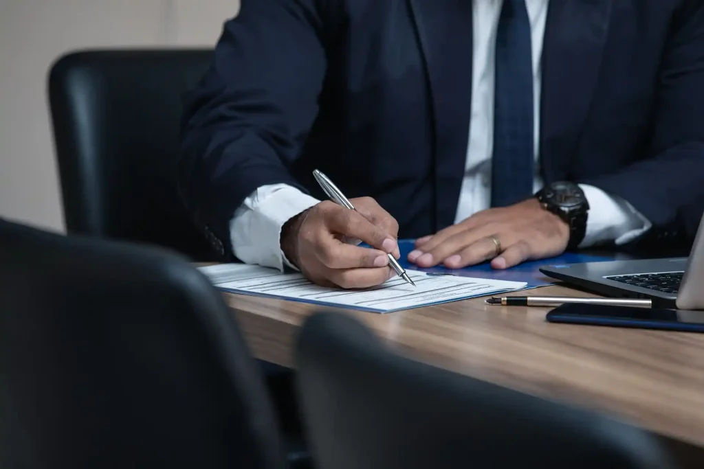 Close-up of a professional man in a suit signing documents on a desk, with a laptop, pen, and clipboard nearby, lemonlawassist