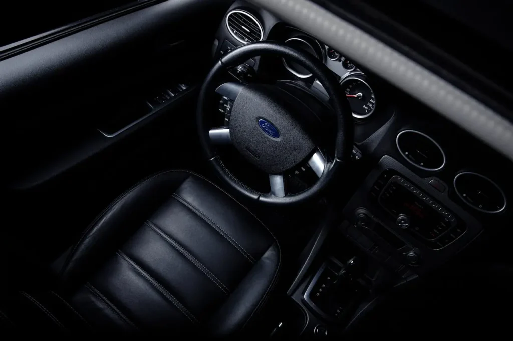 Interior view of a car showcasing the steering wheel with a Ford logo, leather driver's seat, dashboard controls, lemonlawassist