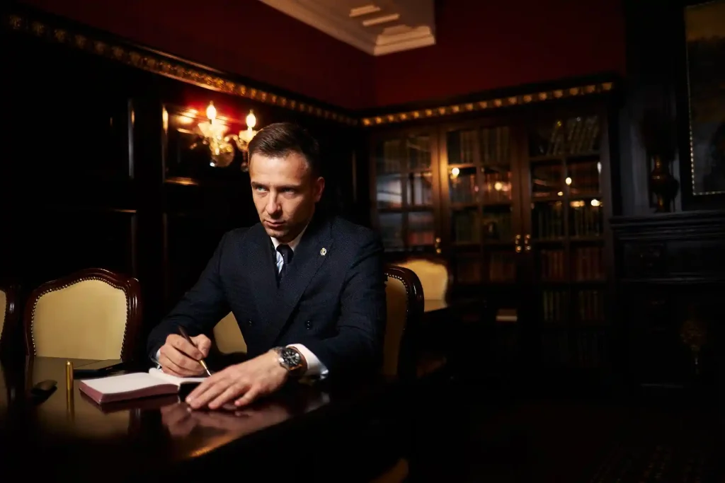 Man in a formal suit sitting at a polished wooden desk in a dimly lit, elegant library, writing in a notebook with bookshelves, lemonlawassist