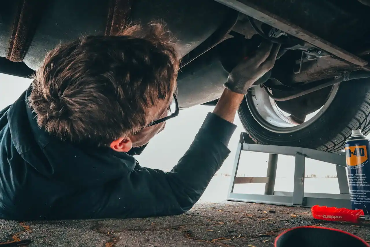 Mechanic lying on the ground under a vehicle, wearing gloves and glasses, working on the car's underside with tools, lemonlawassist