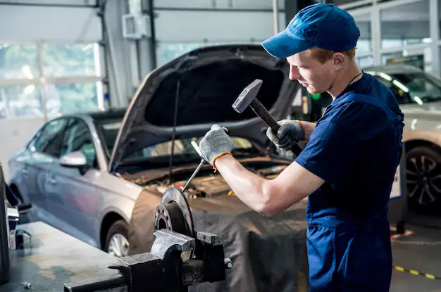 Mechanic wearing a blue uniform and cap working on car brakes with a hammer in an automotive service station, lemonlawassist