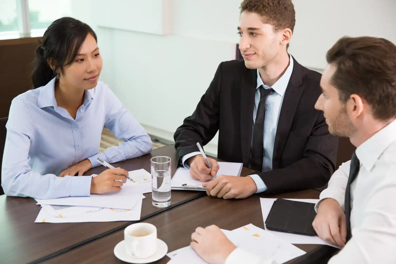 Three lawyers in a meeting, sitting around a table with documents, a tablet, and a cup of coffee, engaged in a discussion, lemonlawassist