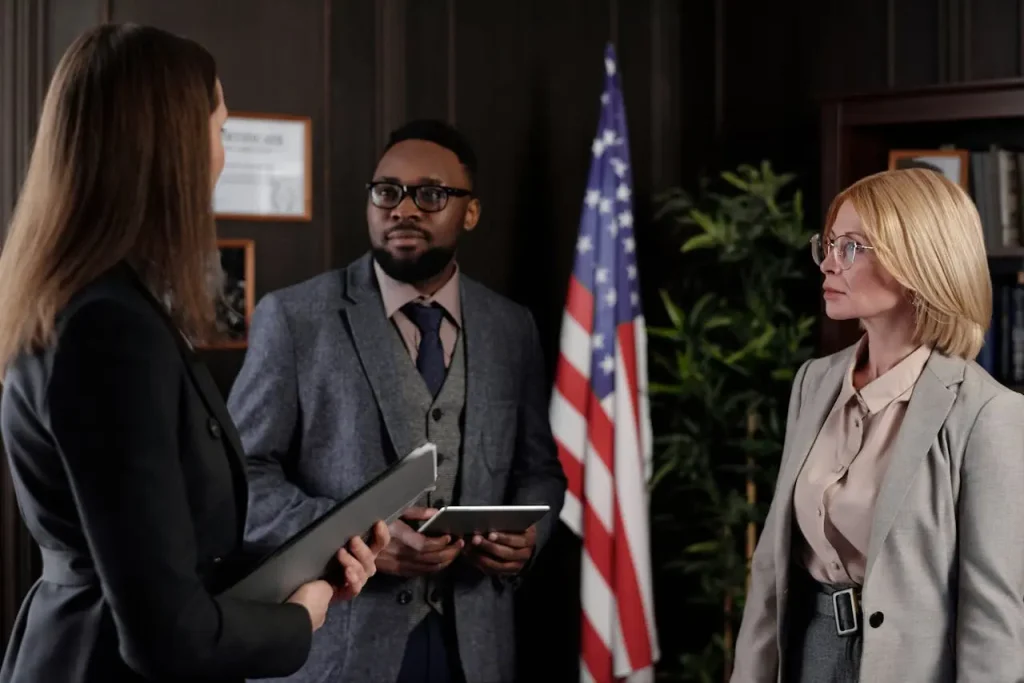 Three professionals in formal attire having a discussion in an office, with an American flag and bookshelves, lemonlawassist
