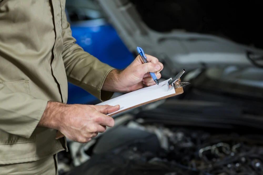 A mechanic in a beige uniform holding a clipboard and pen, writing on a checklist while standing near an open car hood with a blue vehicle in the background, lemonlawassist