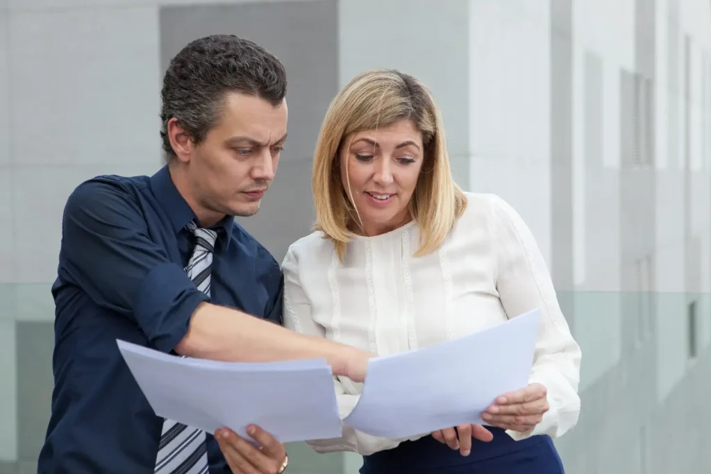 A professional man in a dark shirt and striped tie discusses documents with a smiling woman in a white blouse, lemonlawassist