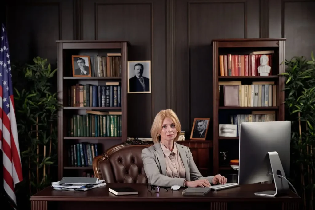 A professional woman sitting at a large wooden desk in a formal office, surrounded by bookshelves, framed photos, lemonlawassist