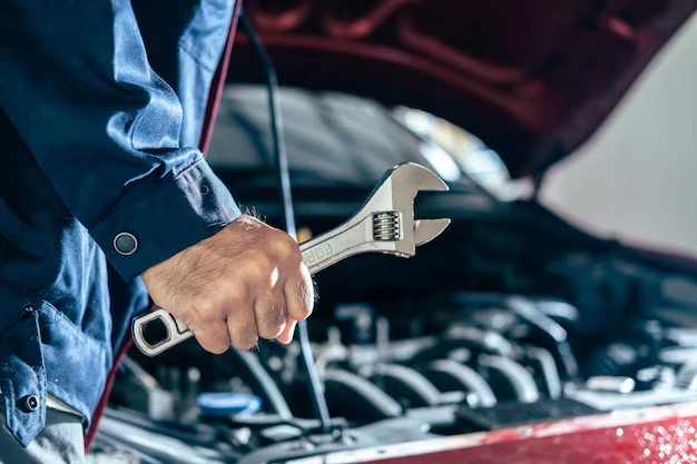Close-up of a mechanic holding a wrench, standing near a car with its hood open, lemonlawassist