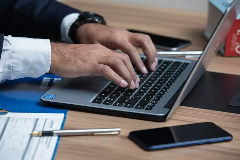 Close-up of a person's hands typing on a laptop keyboard, with a smartphone and documents on a wooden desk, lemonlawassist