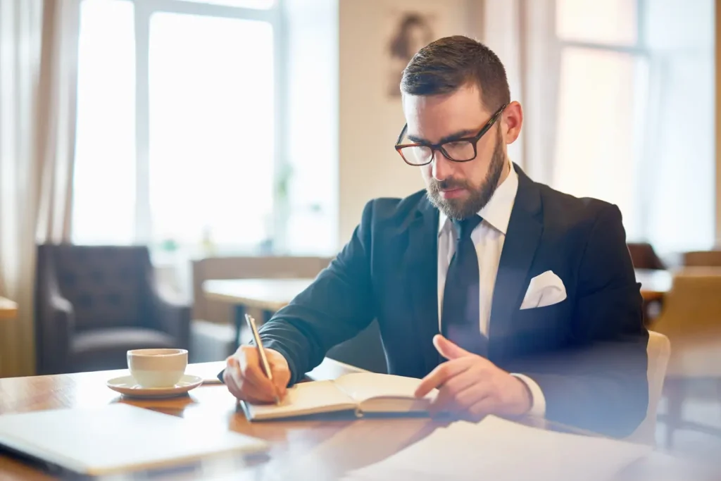 Lawyer in a formal suit writing in a notebook at a desk with a cup of coffee, working in a bright and modern office environment, lemonlawassist