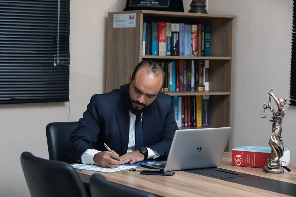 Professional lawyer in a suit writing documents at his desk, with a laptop, legal books, and a Lady Justice statue in an office setting, lemonlawassist