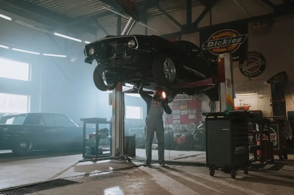A mechanic in overalls working underneath a vintage black car lifted on a hydraulic lift in a well-lit workshop, surrounded by tools and vintage decor, lemonlawassist