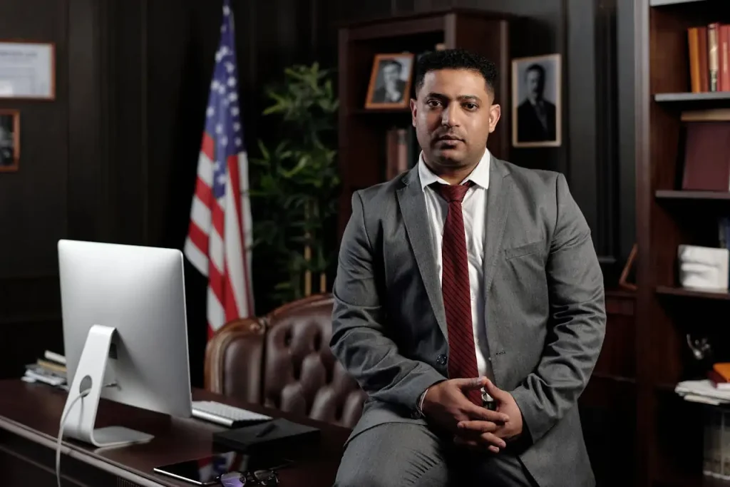 A professional man in a gray suit and red tie sits confidently in a law office, with an American flag, wooden bookshelves, and a desk with a computer in the background, lemonlawassist