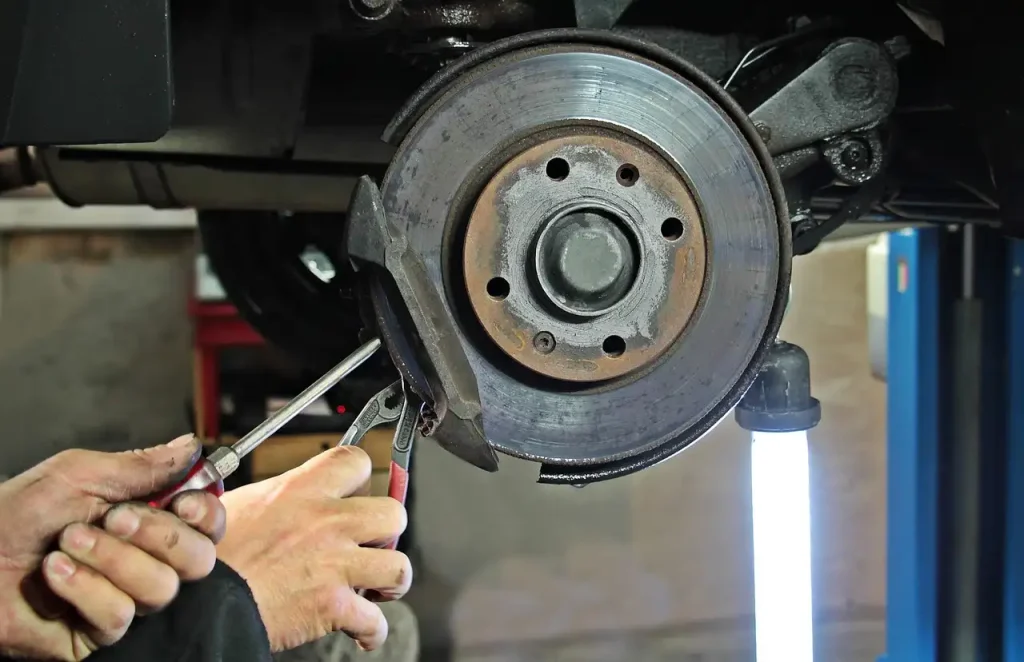 Close-up of a mechanic's hands using tools to repair a car's brake system, with a visible brake disc and caliper under bright workshop lighting, lemonlawassist