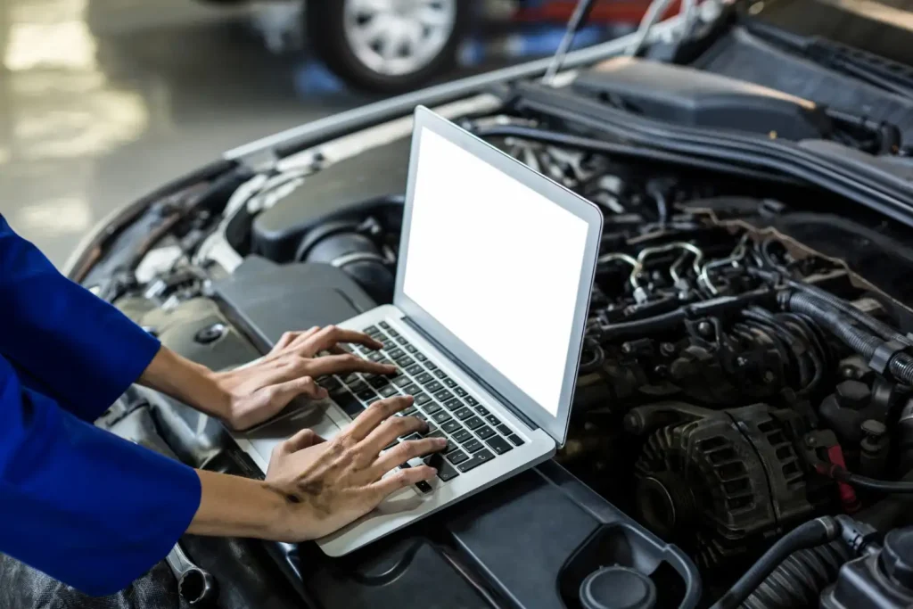 Mechanic in a blue uniform using a laptop while diagnosing a car engine. The laptop rests on the open hood, and the mechanic's hands appear slightly greasy from working on the vehicle, lemonlawassist