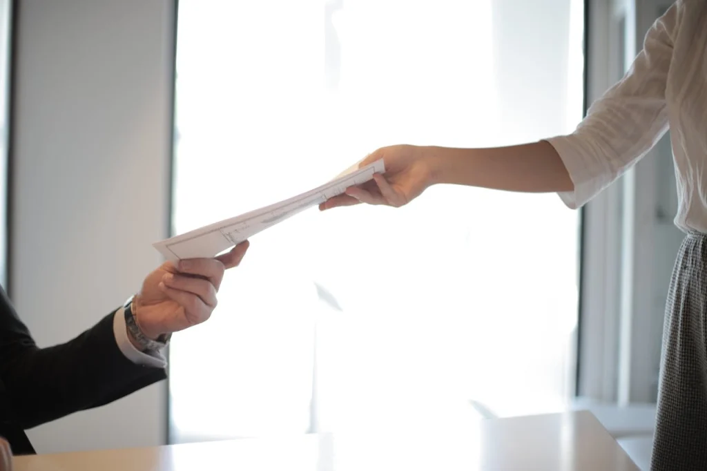 Professional lawyer interaction with a woman handing over a document to a suited individual in an office setting, lemonlawassist