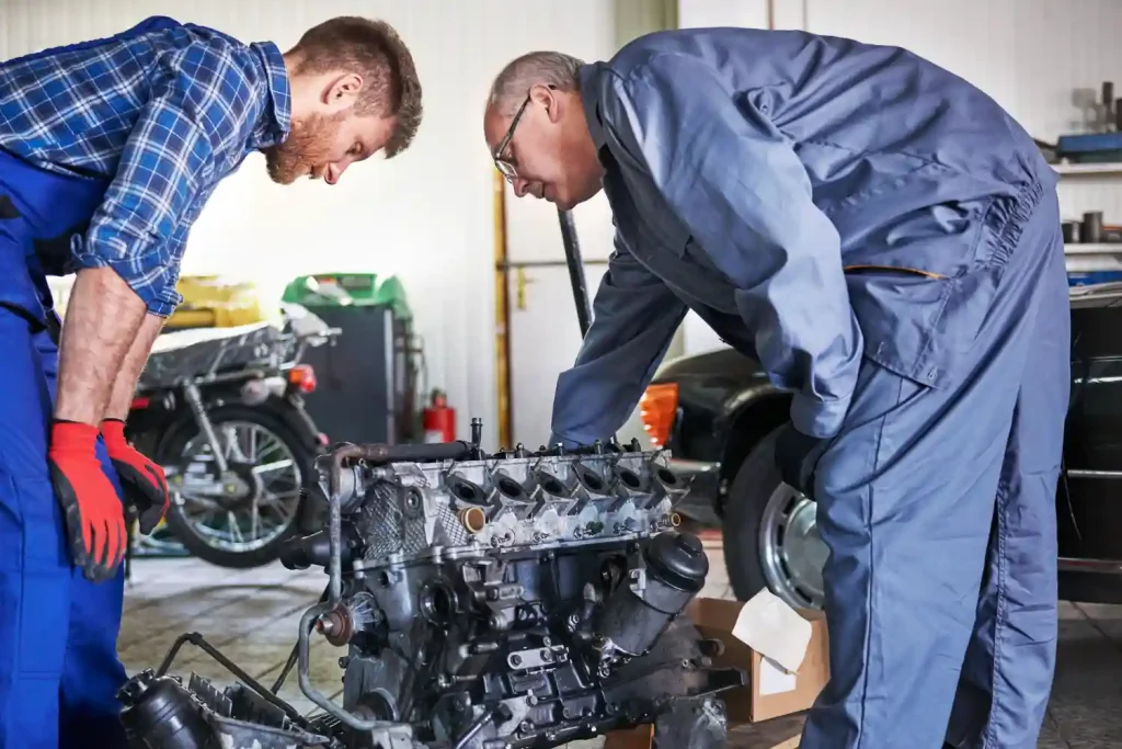 Two mechanics in a workshop examining a disassembled car engine. One wears a blue jumpsuit and leans in closely, while the other, in a plaid shirt and overalls, observes attentively, lemonlawassist