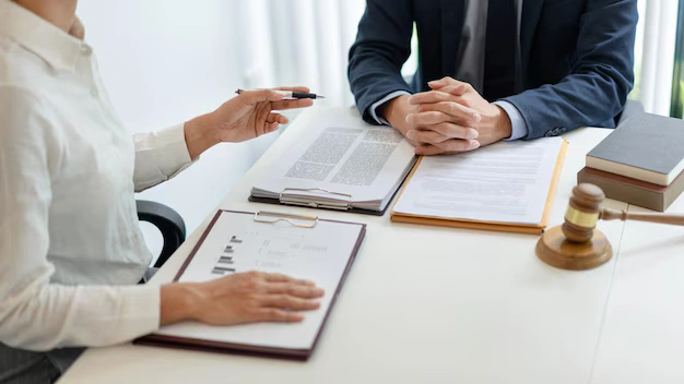 Two professionals lawyers sitting across a desk, reviewing legal documents with a gavel, a stack of books, and paperwork visible in a bright office setting, lemonlawassist