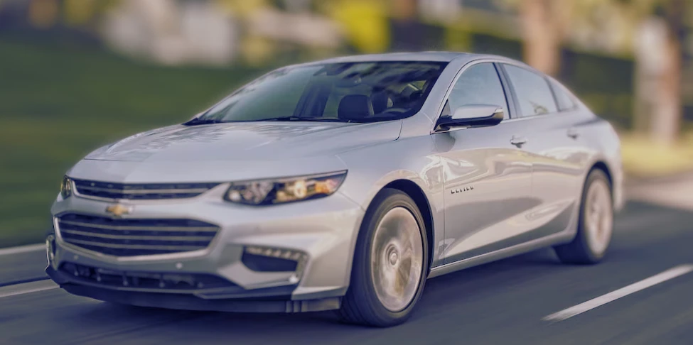 A silver Chevrolet Malibu sedan driving on a suburban road, captured in motion with a blurred background, lemonlawassist