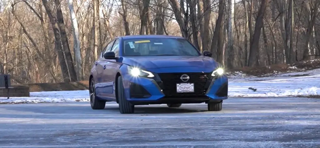 Blue Nissan Altima driving on a snowy, tree-lined road with headlights on during winter, lemonlawassist