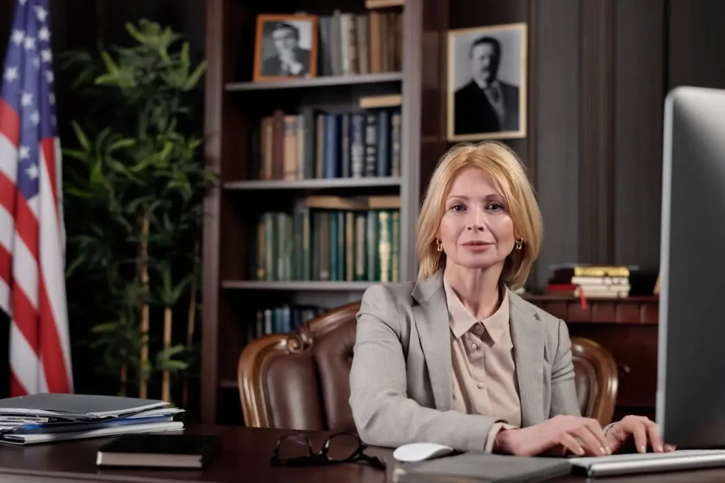 Confident female lawyer in a gray suit sitting at a desk with a computer, books, and documents, set in a professional office, lemonlawassist
