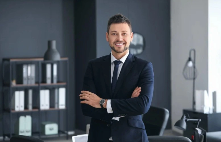 Confident lawyer in a dark suit standing with arms crossed in a modern office, smiling professionally, lemonlawassist
