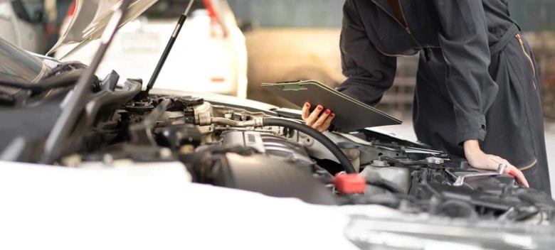 Female auto mechanic in a black uniform inspecting a car engine while holding a clipboard in a well-lit garage, lemonlawassist