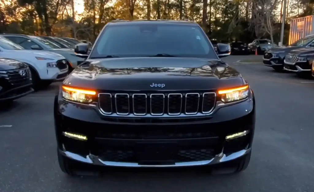Front view of a black Jeep SUV with headlights on, parked in a lot with other vehicles at sunset, lemonlawassist