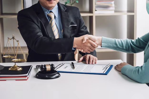 Lawyer in a suit shaking hands with clients over a contract, with a gavel placed on the office desk, symbolizing legal agreement, lemonlawassist