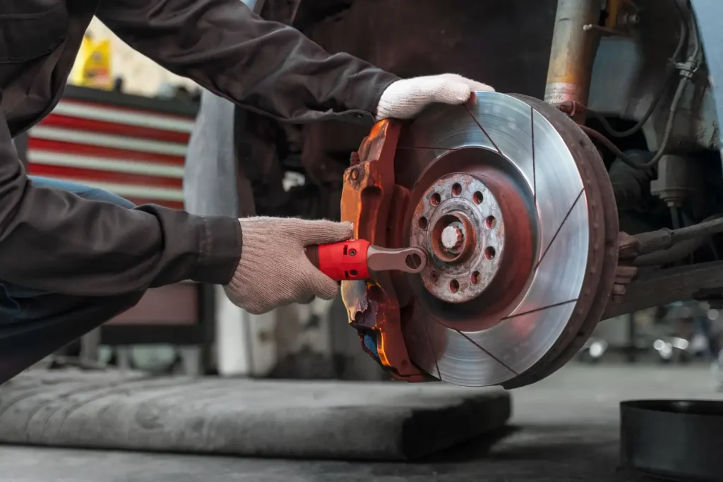 Mechanic wearing gloves using a red ratchet tool to work on a car's brake system, focusing on the brake disc and caliper in an automotive workshop setting, lemonlawassist