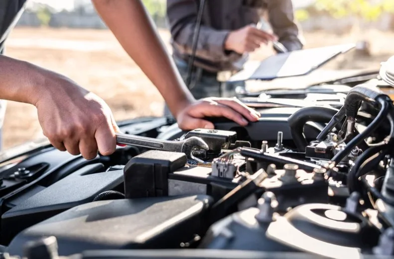 Mechanic working on a car engine outdoors, using a wrench to tighten components while another person takes notes on a clipboard, lemonlawassist
