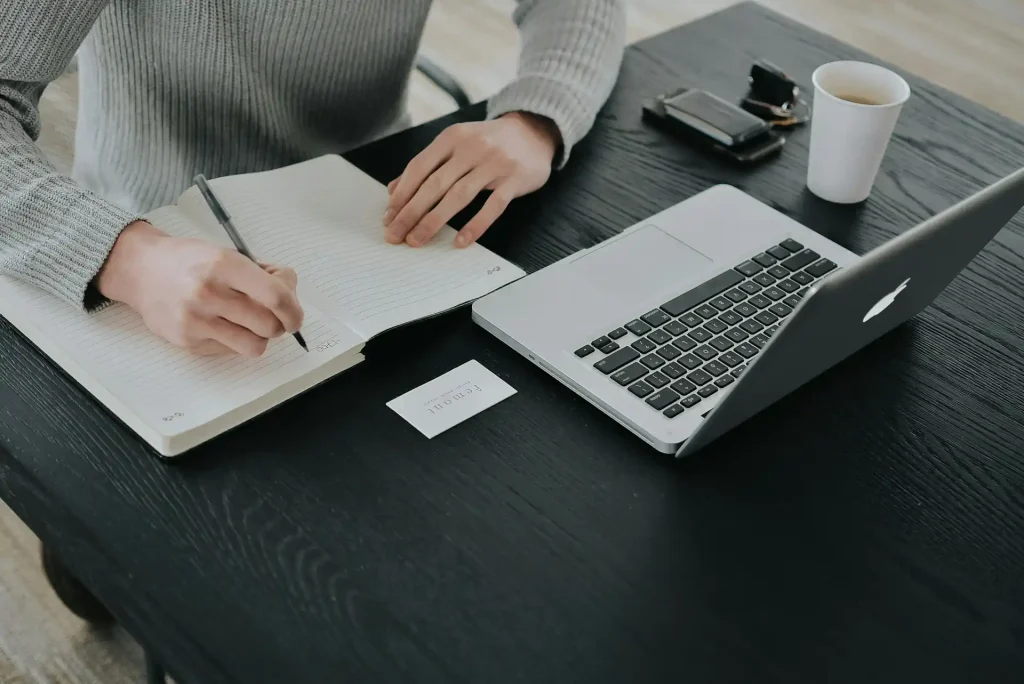Person writing in a notebook with a laptop, coffee cup, and personal items on a black desk, creating a workspace setting, lemonlawassist