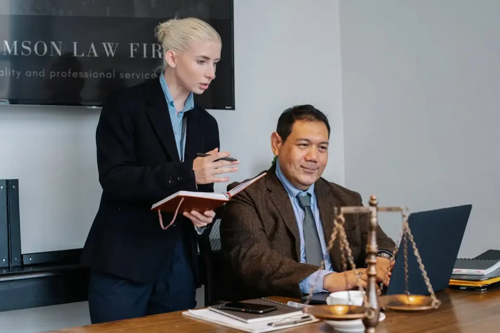 Two professionals in a law office, discussing notes while working on a laptop, with legal scales symbolizing justice on the desk, lemonlawassist