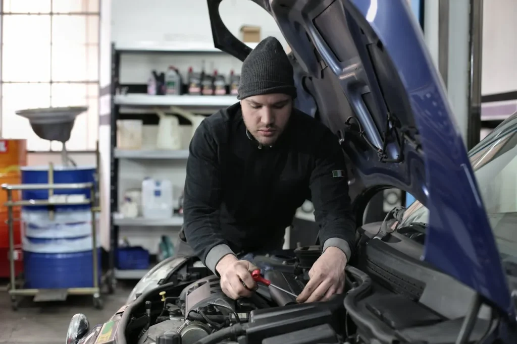 Male mechanic wearing a beanie and black jacket working on a car engine in an auto repair shop, lemonlawassist