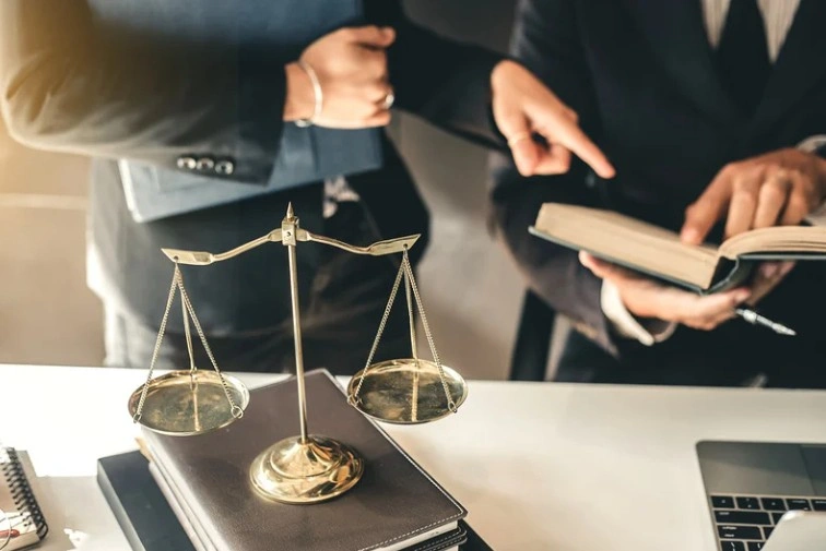 Two legal professionals discussing a case with a book, with a brass balance scale on the desk in the foreground, lemonlawassist