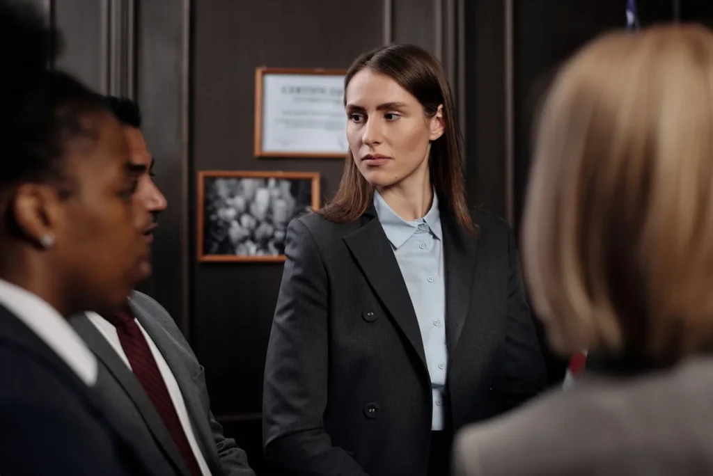 A professional woman wearing a dark suit, observing her colleagues during a business meeting, standing in an office setting.
