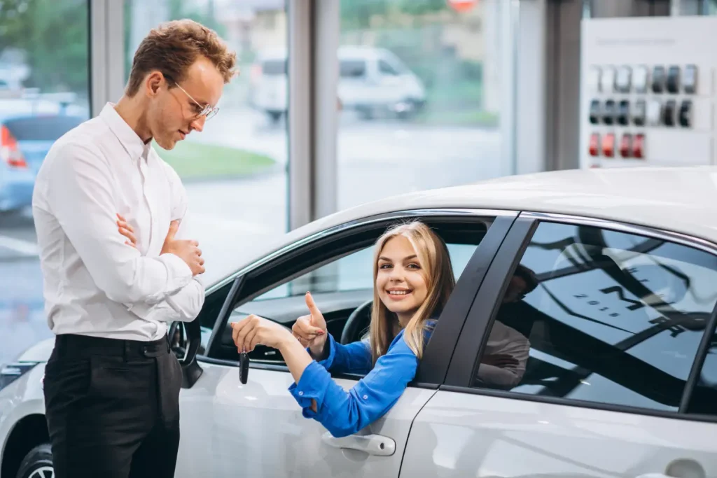 A woman in a blue shirt receiving keys from a salesman in a car dealership while smiling and giving a thumbs up, lemonlawassist